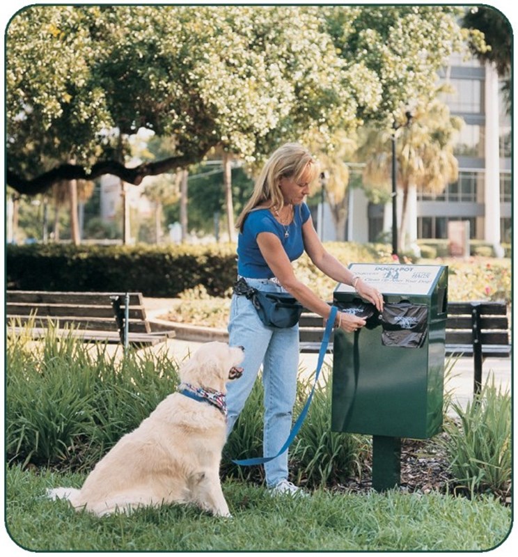 Woman disposing dog waste in green bin at park with golden retriever sitting nearby.