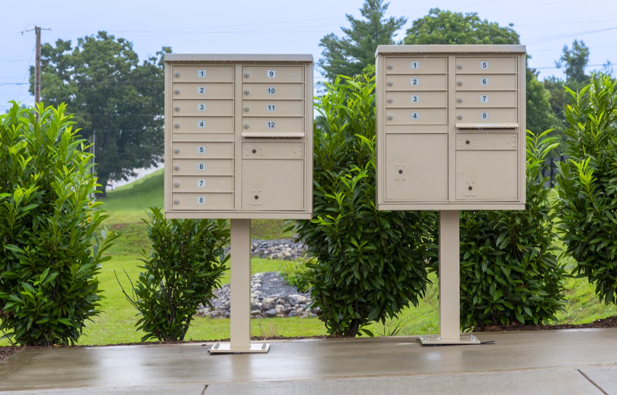 Two beige outdoor mailbox units with numbered compartments on a concrete path, surrounded by greenery.
