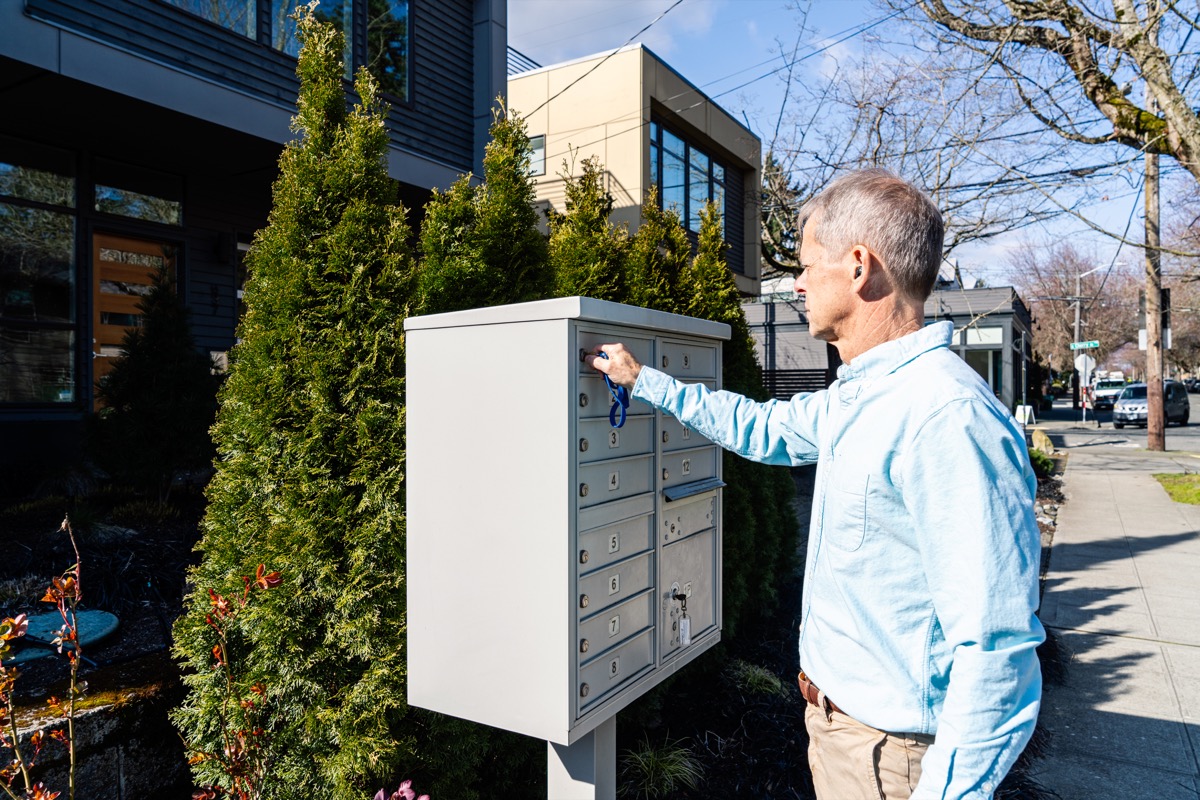 Man in blue shirt unlocking a community mailbox on a sunny residential street.