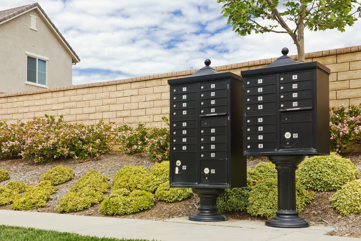 Black community mailboxes on pedestals near a brick wall and landscaped shrubs.