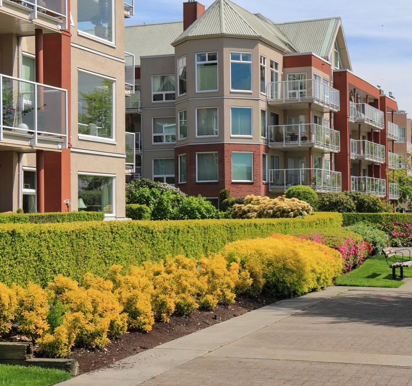 Modern apartment buildings with balconies, surrounded by landscaped gardens and colorful shrubs.